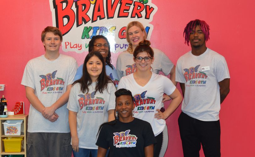 Marcella sitting with the staff of the Bravery Kids Gym in front of a wall with their logo on it
