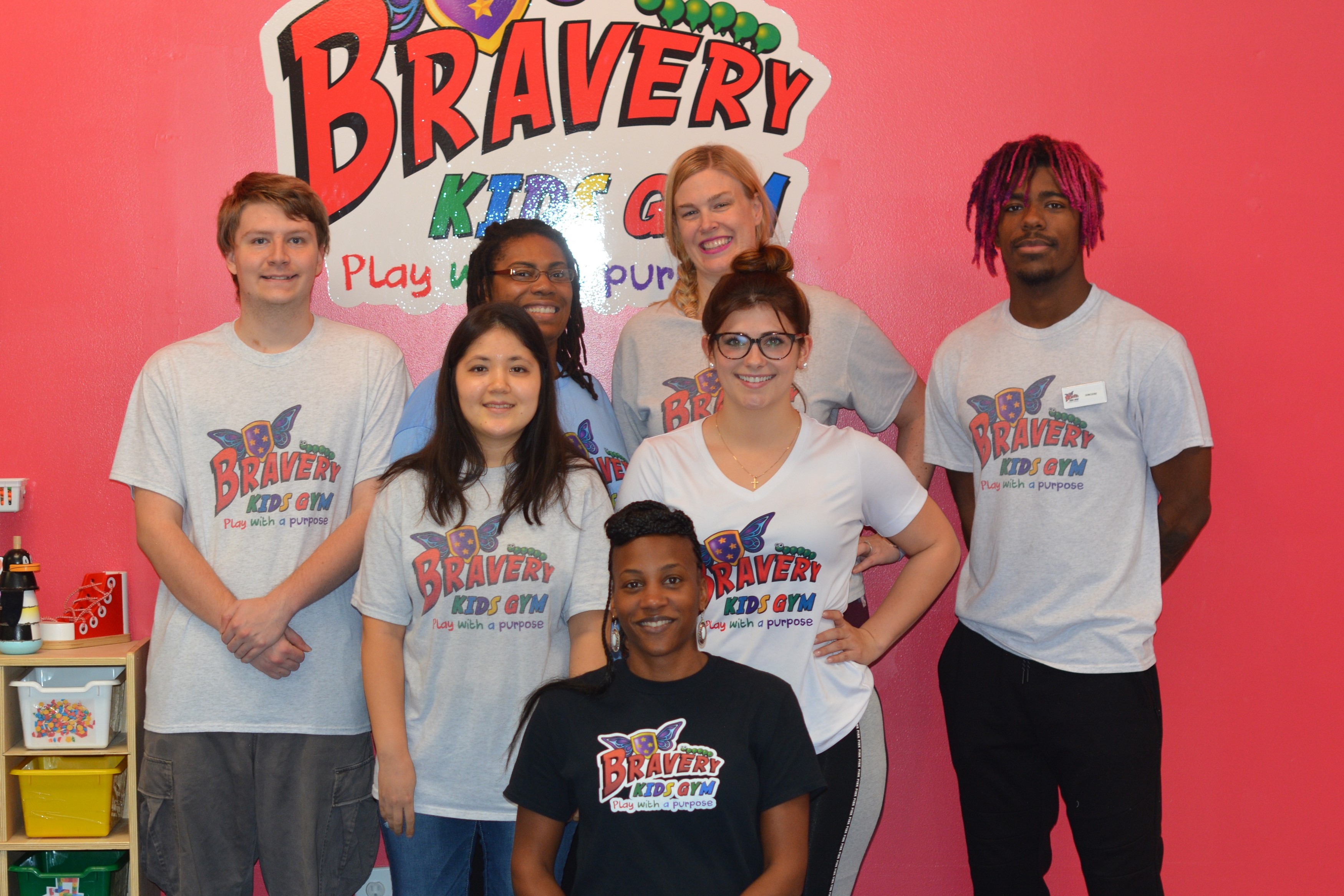 Marcella sitting with the staff of the Bravery Kids Gym in front of a wall with their logo on it