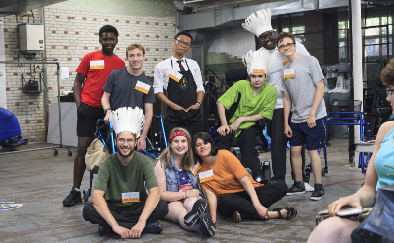 A group of PYD youth and staff gather for a photo backstage. A few are dressed in chef costumes at the 2019 ATT summer show. Three people are sitting on the ground, and six standing.
