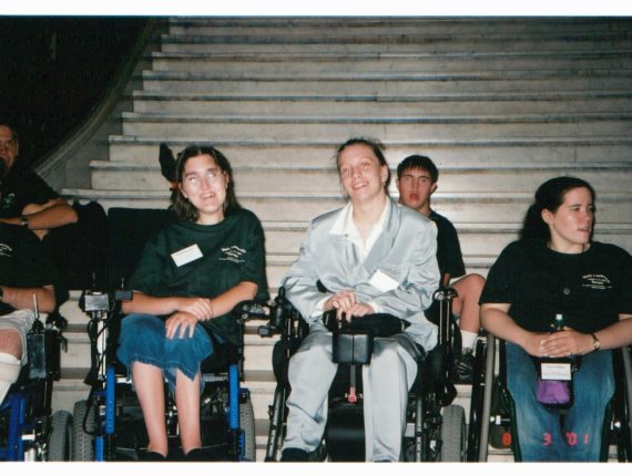 Sarah (left) along with two others smile while sitting in wheel chairs in front of a large white staircase.