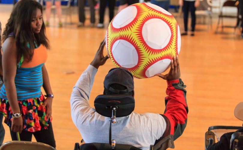 Man in wheel chair is holding up an orange and red ball. Another youth is standing next to them and looking at them.