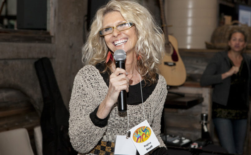 Regina Snowden, a white woman with shoulder length blonde hair and a huge smile, speaking at a microphone