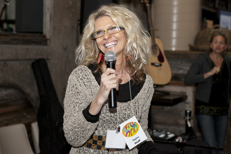 Regina Snowden, a white woman with shoulder length blonde hair and a huge smile, speaking at a microphone