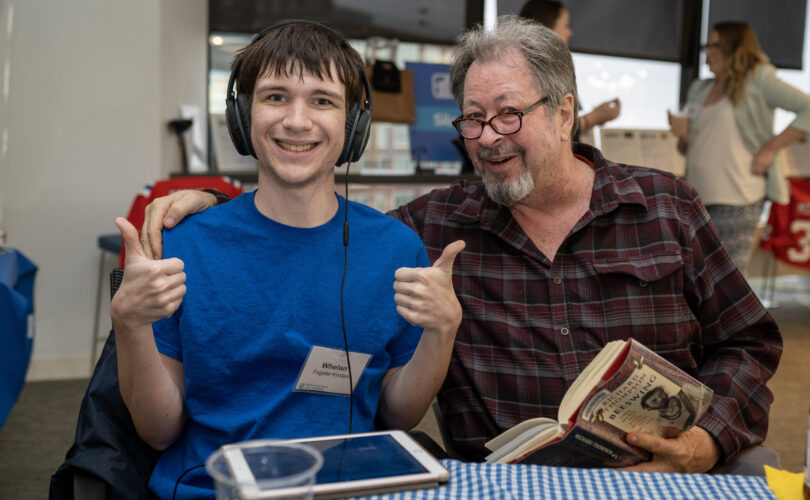 One man smiling and giving thumbs up and another man holding a book and smiling