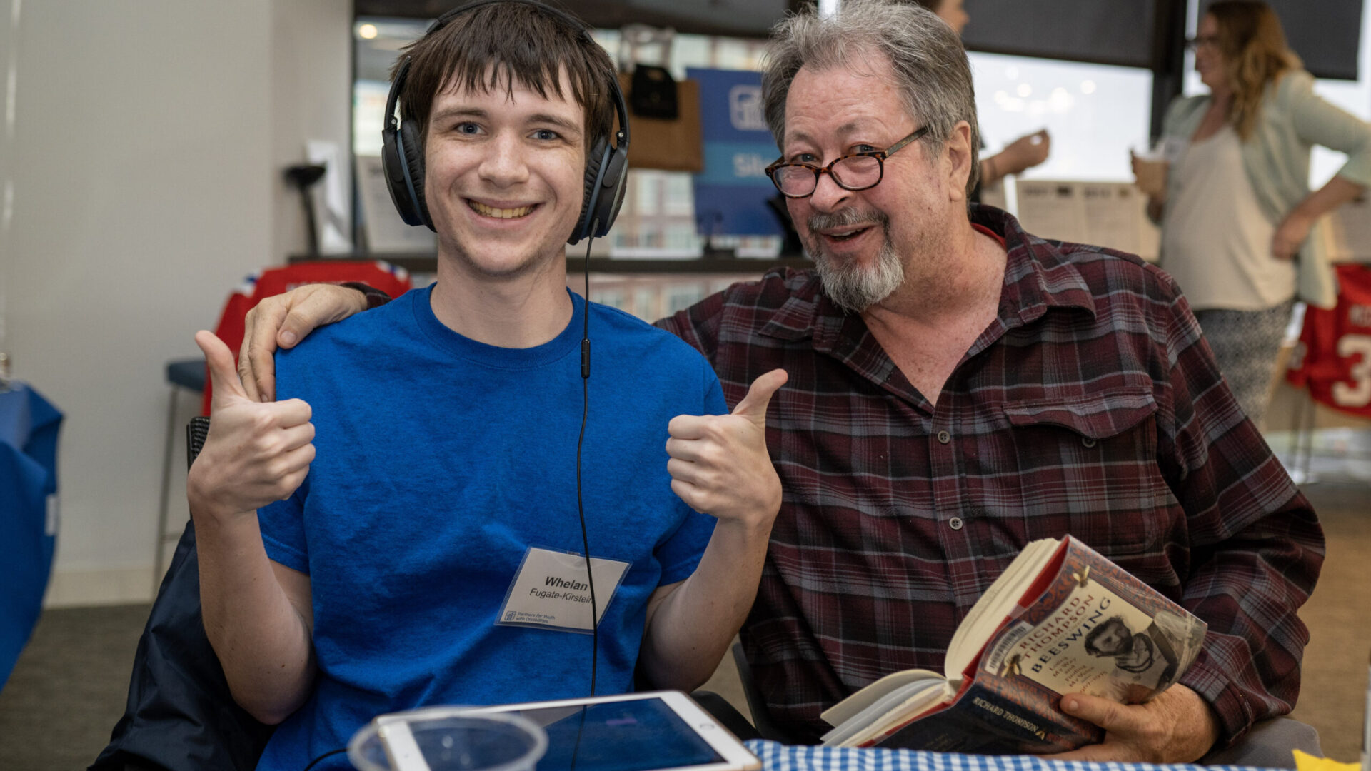 One man smiling and giving thumbs up and another man holding a book and smiling
