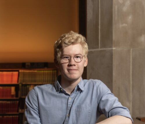 Ansel has curly blond hair and glasses. He is wearing a light blue denim shirt and seated in a chair.