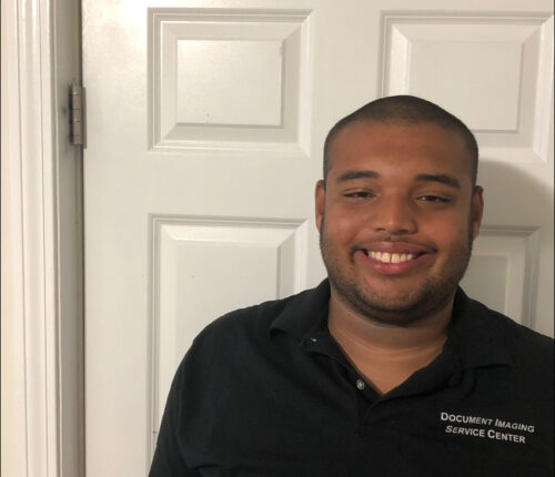 Anthony is wearing a black polo shirt and standing in front of a white door. He has a shaved head and a beard, and is smiling.