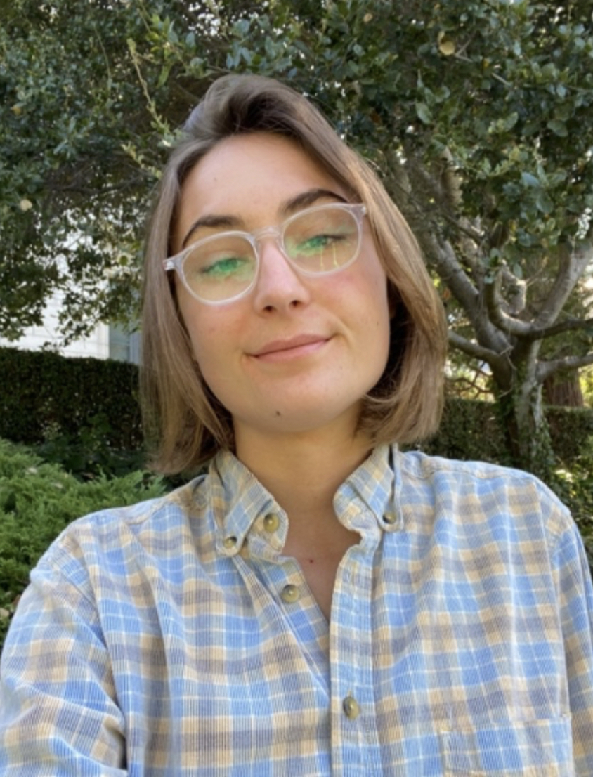 Morgan has medium brown hair cut into a bob and is wearing clear glasses. They are wearing a light blue, white, and yellow plaid button up shirt.