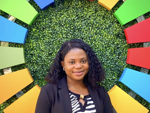 Aanuoluwapo stands in front of a circle of rainbow-colored pieces of wood on a wall of green shrubbery. She is wearing a striped shirt and a black blazer, and has shoulder length black hair.