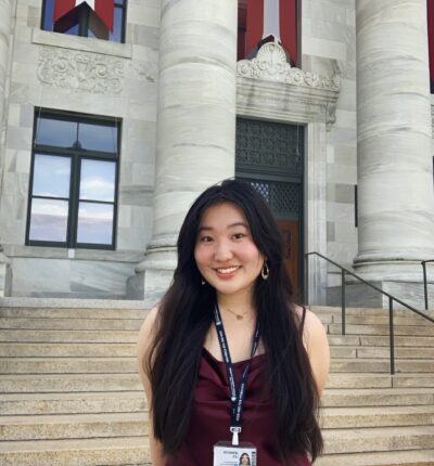 Jennifer has long black hair, and is wearing a maroon colored dress. She is standing in front of the steps and columns of a large building.