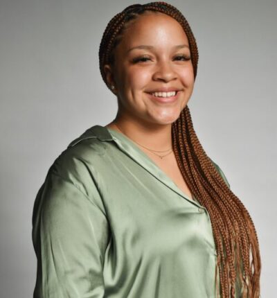 Raven has long brown hair in long braids, and is smiling while wearing a silky light green blouse.